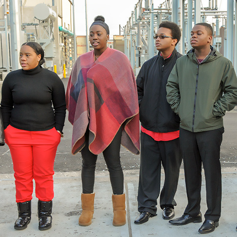 Four singers at a public art launch; industrial equipment in the background